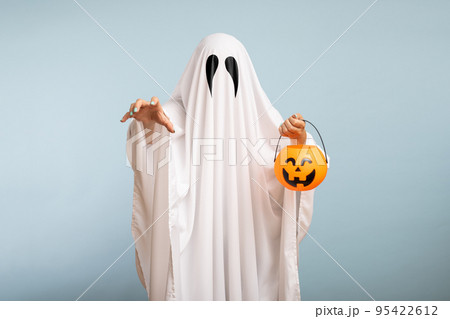 Halloween Concept. A white ghost with black eyes, made from a bedsheet with pumpkin basket for candy on blue background 95422612