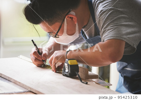 A carpenter measures the planks to assemble the parts and build a wooden table for the customer. A carpenter measures the planks to assemble the parts and build a wooden table for the customer. 95425310
