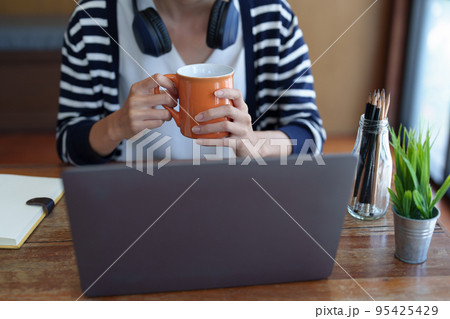 Portrait of a young, attractive Asian teenage girl using a computer and drinking coffee while studying online in the morning at the library. 95425429