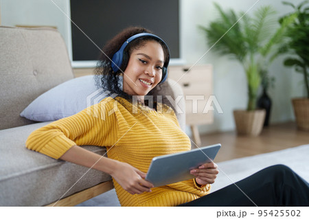 Portrait of an African American with headphones and tablet smiling happily listen music while relaxing on the sofa at home. 95425502