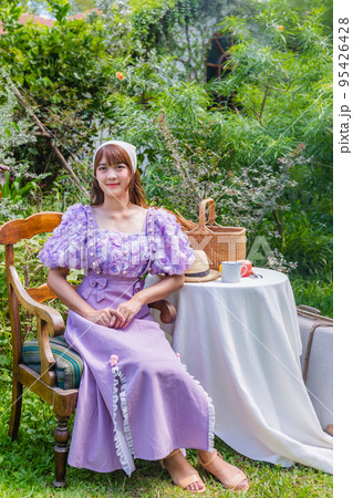 young woman in a princess dress sits drinking tea on a vintage table with cookies and books in a backyard with plants and flowers. 95426428