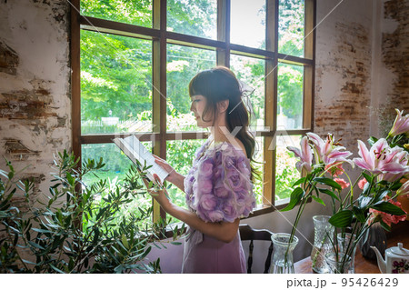 Girl in a princess dress reads a book in a room with windows and lots of green plants and lilies in a vase on the table like a fairy tale. Soft focus Girl in a princess dress reads a book in a room with windows and lots of green plants and lilies in a vase on the table like a fairy tale. Soft focus 95426429