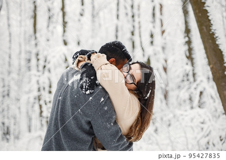 Indian man and caucasian woman walking in winter forest together. Lovely couple hugging. Man wearing grey coat and woman beige coat. 95427835