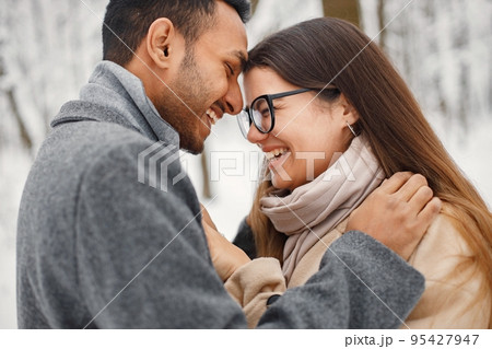 Indian man and caucasian woman walking in winter forest together. Lovely couple hugging. Man wearing grey coat and woman beige coat. Indian man and caucasian woman walking in winter forest together. Lovely couple hugging. Man wearing grey coat and woman beige coat. 95427947