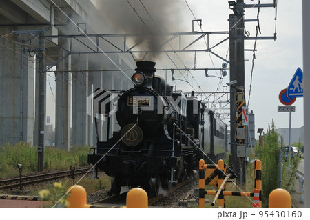 勢いよく煙を噴き上げながら踏切に近づく蒸気機関車の景色 95430160