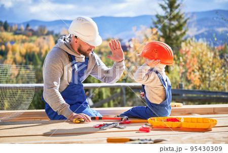 Father with toddler son building wooden frame house. Male builder giving high five to kid on construction site, wearing helmet and blue overalls on sunny day. Carpentry and family concept. 95430309