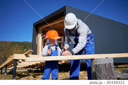 Father with toddler son building wooden frame house. Male builders hammering nail into plank on construction site, wearing helmet and blue overalls on sunny day. Carpentry and family concept. Father with toddler son building wooden frame house. Male builders hammering nail into plank on construction site, wearing helmet and blue overalls on sunny day. Carpentry and family concept. 95430321
