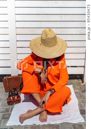 Stylish woman in an orange suit with a hat sits on a rug on a white striped background. On the hands are jewelry rings and bracelets, sandals and a bag stand side by side. Stylish woman in an orange suit with a hat sits on a rug on a white striped background. On the hands are jewelry rings and bracelets, sandals and a bag stand side by side. 95430473