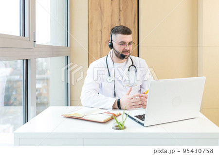 Young doctor man in headphones and with a microphone, advises on video communication of the patient, sits at the table, uses a laptop Young doctor man in headphones and with a microphone, advises on video communication of the patient, sits at the table, uses a laptop 95430758