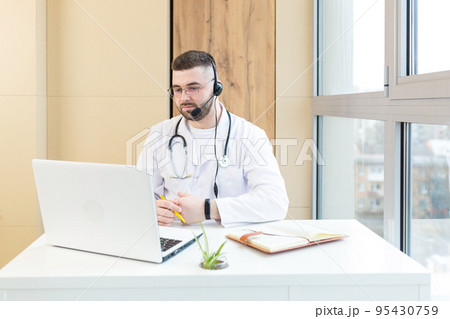 Young doctor man in headphones and with a microphone, advises on video communication of the patient, sits at the table, uses a laptop Young doctor man in headphones and with a microphone, advises on video communication of the patient, sits at the table, uses a laptop 95430759