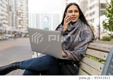 female freelancer speaks on a mobile phone and works on a laptop Sitting on a bench against the female freelancer speaks on a mobile phone and works on a laptop Sitting on a bench against the 95431921