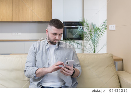 Young man at home sitting on the couch, using the phone, talking on the phone, talking on video Young man at home sitting on the couch, using the phone, talking on the phone, talking on video 95432012