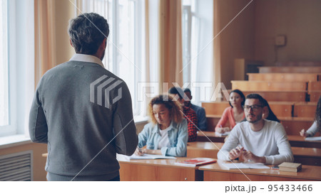Bearded mature man professor is reading lecture talking and gesturing while students are listening and writing sitting at tables in spacious university classroom. 95433466
