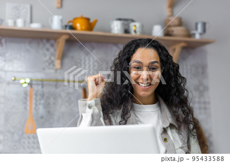 Close up photo of happy and smiling woman looking at laptop screen at home rejoicing and celebrating victory triumph holding hand up, Hispanic woman with curly hair and glasses using netbook. 95433588