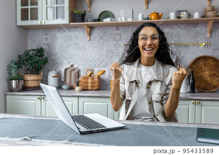 Portrait of successful woman at home, Hispanic woman looking at camera and happy celebrating victory triumph, businesswoman using laptop for remote work, freelancer with curly hair and glasses. 95433589