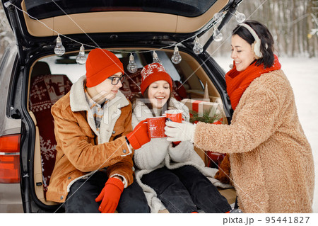 Mother, teens brother and sister spending time together in the winter park. Family sitting in open car's trunk and holding red cups with tea. Family wearing warm jackets and knitted hats. Mother, teens brother and sister spending time together in the winter park. Family sitting in open car's trunk and holding red cups with tea. Family wearing warm jackets and knitted hats. 95441827