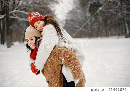 Joyful girl and her beautiful mother spending time together in the winter park. Woman wearing warm jacket and red sweater. Mother carrying her daughter piggyback. 95441872