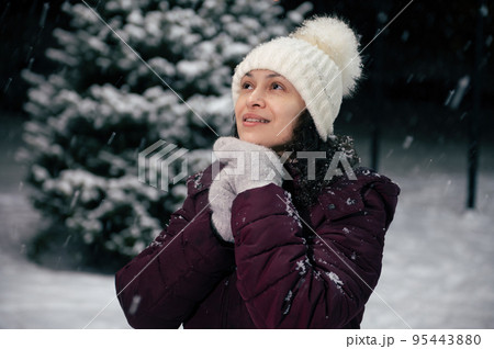 Close-up portrait of charming dark-haired multi-ethnic woman in warm clothes, white wool hat and mittens in the snow covered nature. Snowfall. Winter holidays. Happy New Year. Mmerry Christmas concept Close-up portrait of charming dark-haired multi-ethnic woman in warm clothes, white wool hat and mittens in the snow covered nature. Snowfall. Winter holidays. Happy New Year. Mmerry Christmas concept 95443880