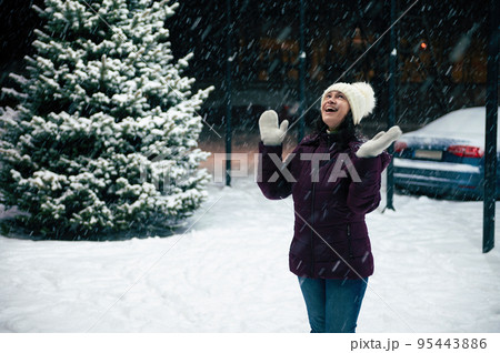 Joyful Latin American woman in warm clothes, enjoys the snowfall, rejoices in the first snow, standing on a snowy street illuminated by New Year's garlands. Merry Christmas and happy winter holidays 95443886