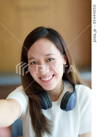 A portrait of a young Asian woman wearing headphones showing a smiling face taking a selfie. 95444050