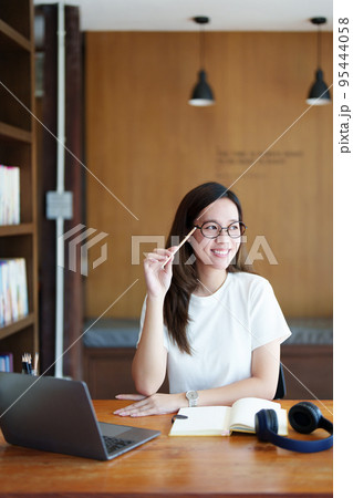 Portrait of a teenage Asian woman using a computer and notebook to study online via video conferencing on a wooden desk in library. 95444058