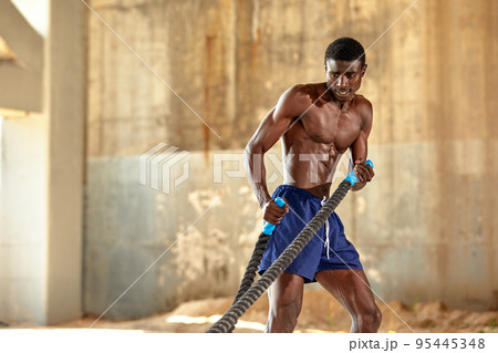 Rope workout. Sport man doing battle ropes exercise outdoor. Black male athlete exercising, doing functional fitness training with heavy rope 95445348