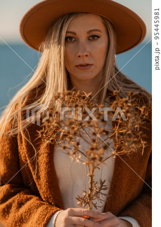 A woman walking along the coast near the sea. An elegant lady in a brown coat and a hat with fashionable makeup walks on the seashore 95445981