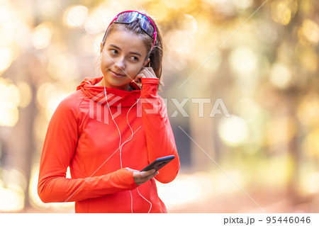 Young female athlete in sportswear listens to music on smartphone and puts headphones before running Young female athlete in sportswear listens to music on smartphone and puts headphones before running 95446046