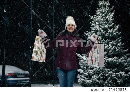 Beautiful African American woman in warm clothes, wool hat and mittens, with shopping bags in her hands, smiling a toothy smile, looking at the camera, on a snowy winter evening. Enjoying winter 95446653
