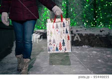 Selective focus on shopping bags with Christmas pattern in the hands of a pretty woman, walking down the snow covered city street, on a winter night, against fir-trees illuminated by festive garlands 95446655