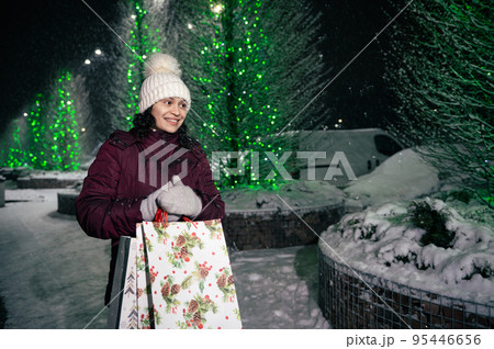 Charming multi-ethnic woman with shopping bags, walking down the city street, lightened by festive illumination at winter snowy night. Christmas holidays. Enjoying winter. Fir-trees with garlands 95446656