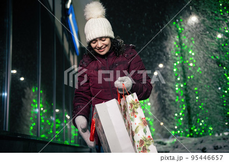 Charming multi-ethnic woman with shopping bags and Christmas gifts, walking down the street, lightened by festive illumination at winter snowy night. Christmas holidays. Enjoying winter 95446657