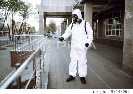 Man in protective suit and mask sprays disinfector onto the railing in the public place. Covid -19. 95447065