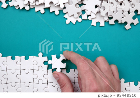 The hand folds a white jigsaw puzzle and a pile of uncombed puzzle pieces lies against the background of the blue surface. Texture photo with space for text 95448510