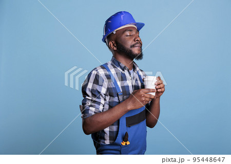 Relaxed construction worker enjoying a cup of favorite coffee before starting work on an interior renovation. Builder wearing coveralls and hard hat against blue background in studio shot. Relaxed construction worker enjoying a cup of favorite coffee before starting work on an interior renovation. Builder wearing coveralls and hard hat against blue background in studio shot. 95448647