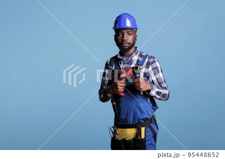 Professional construction worker with coveralls and work tools against blue background. Studio shot african american man looking at camera holding electric cordless drill. Professional construction worker with coveralls and work tools against blue background. Studio shot african american man looking at camera holding electric cordless drill. 95448652