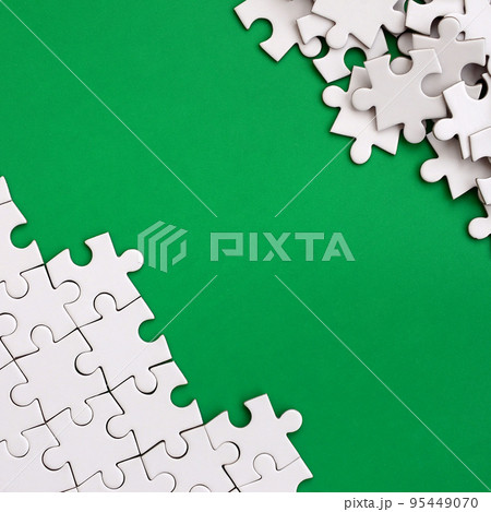 Fragment of a folded white jigsaw puzzle and a pile of uncombed puzzle elements against the background of a green surface. Texture photo with space for text 95449070