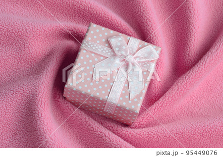 A small gift box in pink with a small bow lies on a blanket of soft and furry light pink fleece fabric with a lot of relief folds. Packing for a gift to your lovely girlfriend A small gift box in pink with a small bow lies on a blanket of soft and furry light pink fleece fabric with a lot of relief folds. Packing for a gift to your lovely girlfriend 95449076