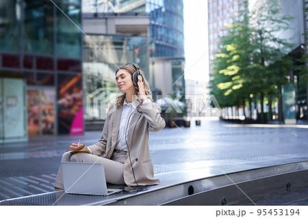 Smiling businesswoman relaxing, listetning to music in headphones from her laptop, sitting alone on street of city centre, wearing suit 95453194