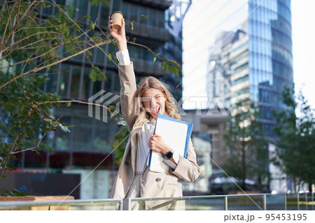 Enthusiastic businesswoman feels upbeat after morning cup of coffee, raise her cup up and dances outside 95453195