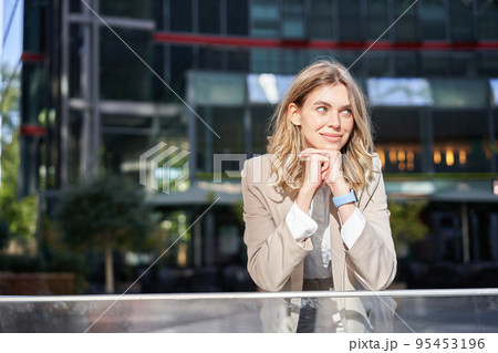 Beautiful young woman in beige suit, standing near office buildings in city center, smiling and looking dreamy 95453196