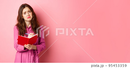 Sad young woman writing her feelings in diary, looking upset and gloomy, feeling heartbroken, sharing memories in planner, standing against pink background 95453359