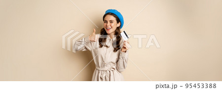 Concept of shopping and sale. Happy young woman in stylish clothes, showing credit card store discount and thumbs up, recommending, standing over beige background Concept of shopping and sale. Happy young woman in stylish clothes, showing credit card store discount and thumbs up, recommending, standing over beige background 95453388