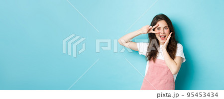 Portrait of excited attractive woman showing v-sign near eye, making kawaii gesture and smiling happy at camera, standing upbeat against blue background Portrait of excited attractive woman showing v-sign near eye, making kawaii gesture and smiling happy at camera, standing upbeat against blue background 95453434