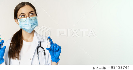 Covid-19 vaccination and healthcare concept. Young doctor in medical mask showing syringe and vaccine from coronavirus omicron variant, standing over white background Covid-19 vaccination and healthcare concept. Young doctor in medical mask showing syringe and vaccine from coronavirus omicron variant, standing over white background 95453744