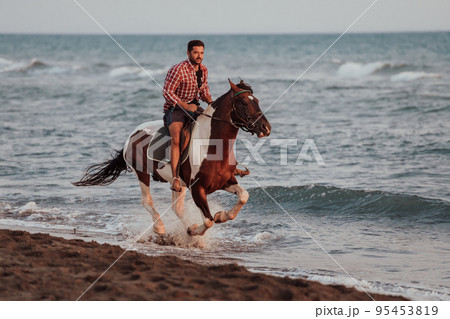 A modern man in summer clothes enjoys riding a horse on a beautiful sandy beach at sunset. Selective focus A modern man in summer clothes enjoys riding a horse on a beautiful sandy beach at sunset. Selective focus 95453819
