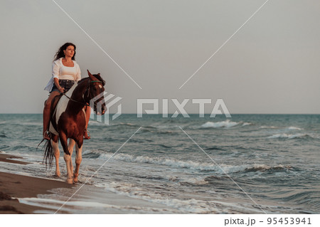 Woman in summer clothes enjoys riding a horse on a beautiful sandy beach at sunset. Selective focus Woman in summer clothes enjoys riding a horse on a beautiful sandy beach at sunset. Selective focus 95453941