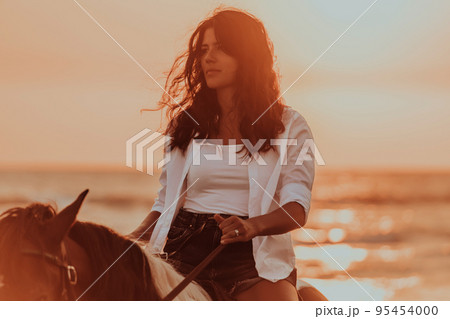 Woman in summer clothes enjoys riding a horse on a beautiful sandy beach at sunset. Selective focus Woman in summer clothes enjoys riding a horse on a beautiful sandy beach at sunset. Selective focus 95454000
