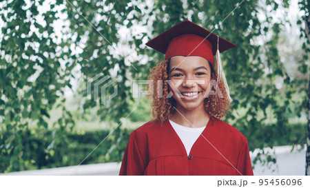 Portrait of pretty African American girl happy graduating student in red gown and mortarboard standing outdoors, smiling and looking at camera. Youth and education concept. Portrait of pretty African American girl happy graduating student in red gown and mortarboard standing outdoors, smiling and looking at camera. Youth and education concept. 95456096