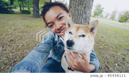 Point of view shot of cheerful African American teenage girl taking selfie with adorable shiba inu dog holding camera, posing and looking at camera. People, nature and animals concept. Point of view shot of cheerful African American teenage girl taking selfie with adorable shiba inu dog holding camera, posing and looking at camera. People, nature and animals concept. 95456111